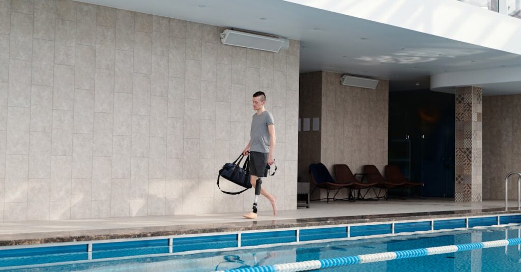 Man with prosthetic leg walking beside an indoor swimming pool carrying a gym bag.