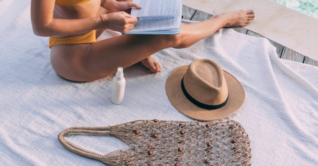 Adult female in swimwear relaxing by the pool, reading with a summer hat and bag nearby.