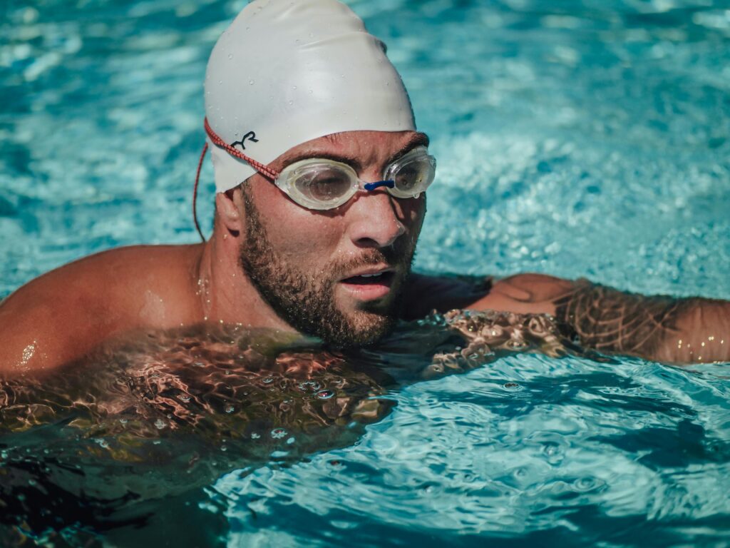 A focused male swimmer with tattoos and beard wearing goggles and a swimming cap in a clear blue pool.